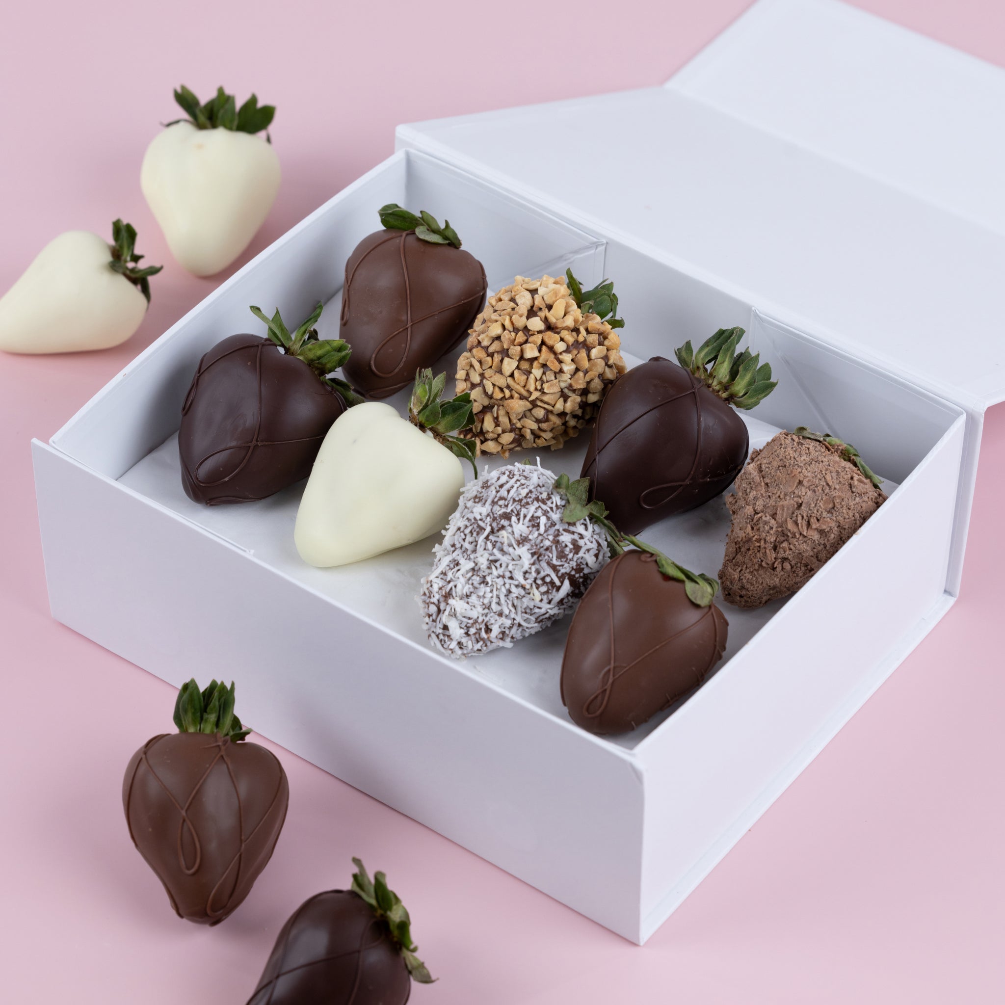 A white gift box with an assortment of chocolate dipped strawberries with swirl patterns, peanut coating and coconut flakes