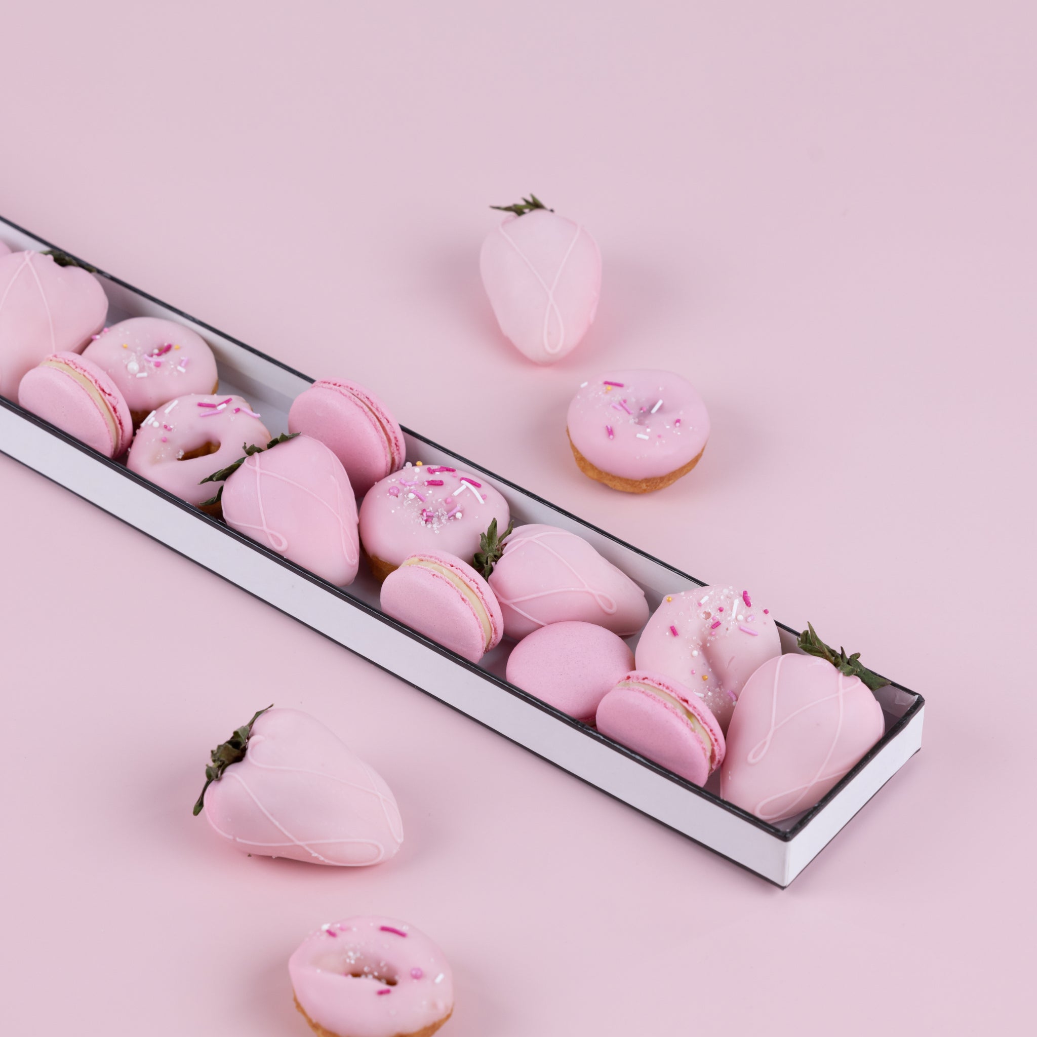 A long, white tray of pink mini doughnuts adorned with sprinkles, pink macarons and pink chocolate dipped strawberries