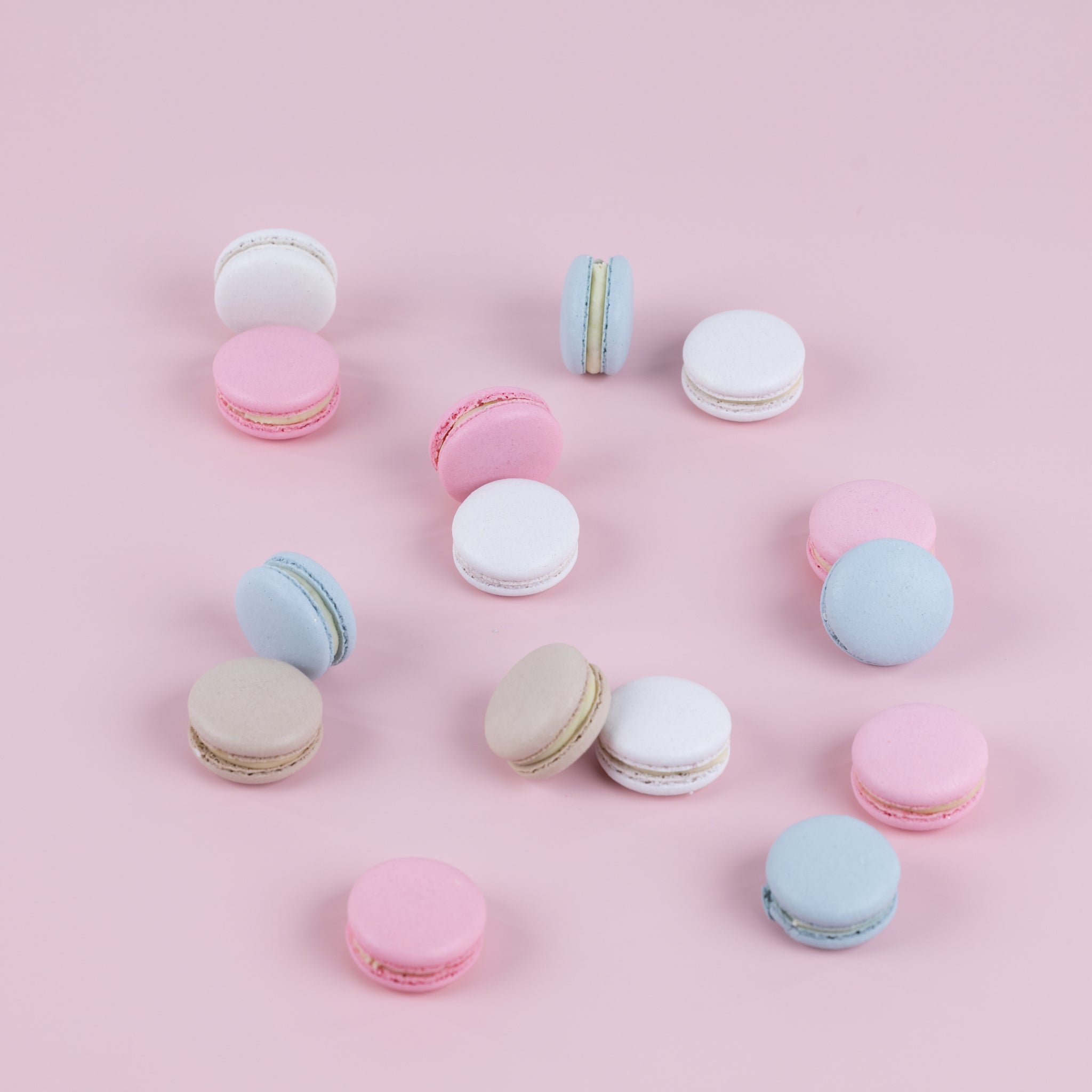 Handmade macarons coloured in white, pink, baby blue and champagne, scattered against a soft pink background