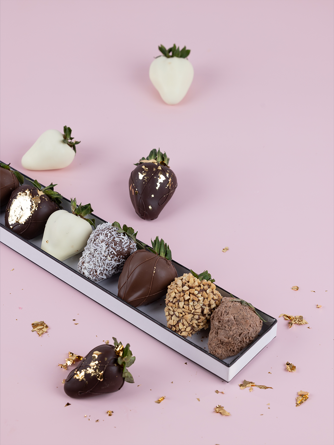 Assortment of chocolate-dipped strawberries with gold leaf, chocolate flakes and coconut arranged on a soft pink backdrop