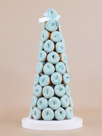 A tower made of baby blue, chocolate coated doughnuts with a designer sprinkle arrangement and a blue ribbon placed on top