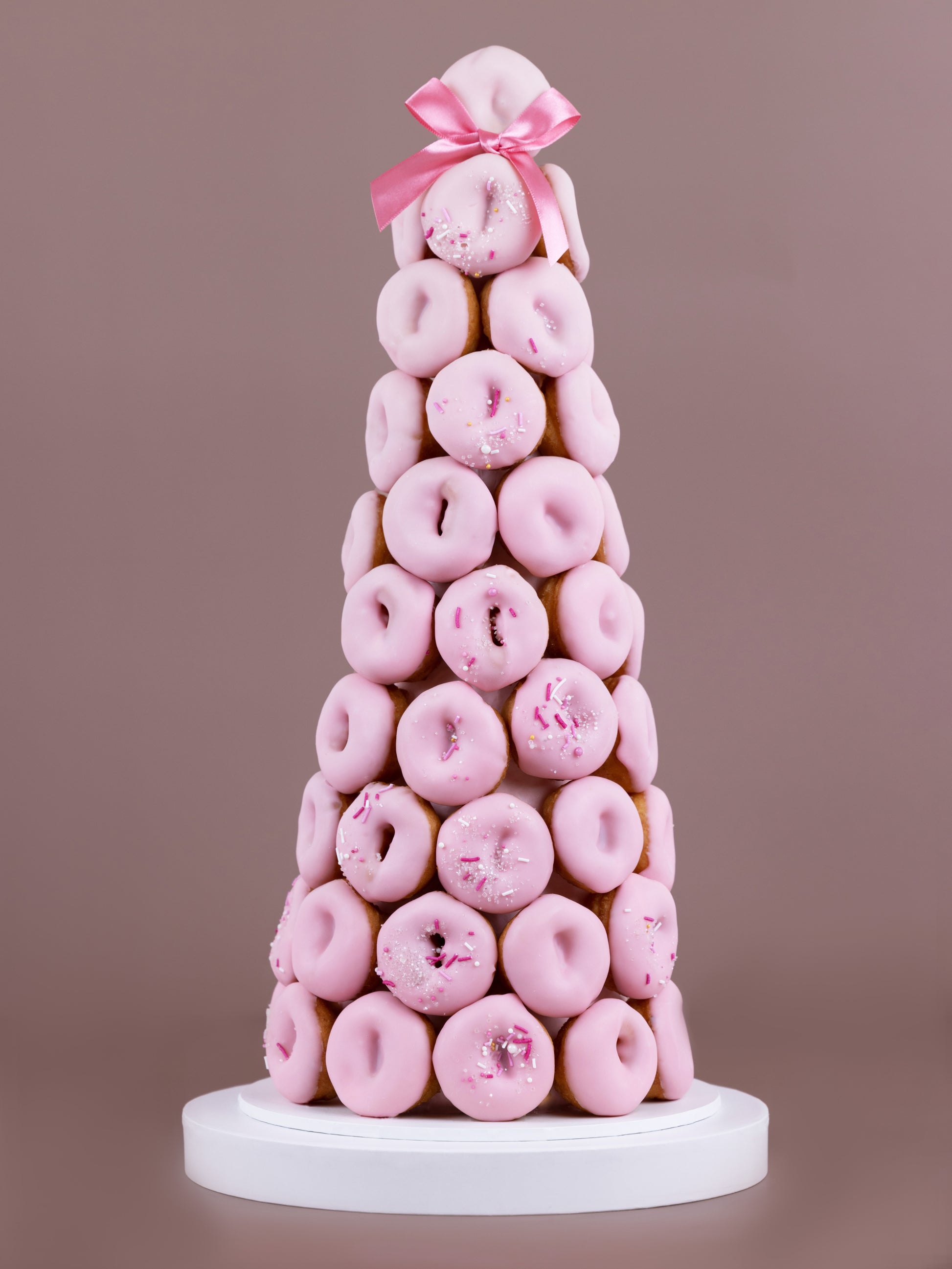 A tower made of pink, chocolate coated doughnuts with a designer sprinkle arrangement and a pink ribbon placed on top